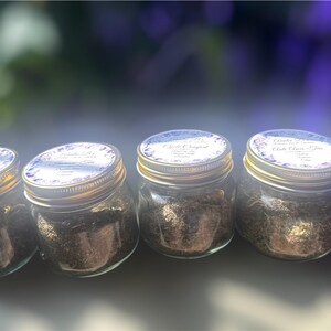 Puede incluir: Cuatro peque&ntilde;os frascos de vidrio con tapas doradas, llenos de una sustancia oscura y granulada. Cada frasco tiene una etiqueta blanca con detalles florales morados. Los frascos est&aacute;n dispuestos en fila, ligeramente inclinados.