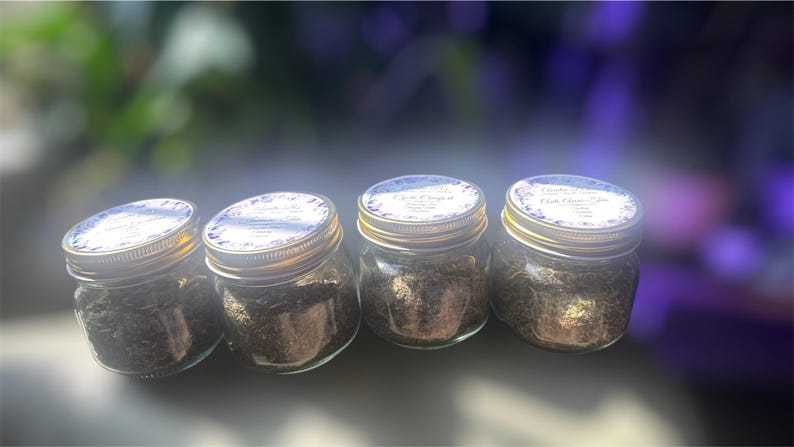 Puede incluir: Cuatro peque&ntilde;os frascos de vidrio con tapas doradas, llenos de una sustancia oscura y granulada. Cada frasco tiene una etiqueta blanca con detalles florales morados. Los frascos est&aacute;n dispuestos en fila, ligeramente inclinados.