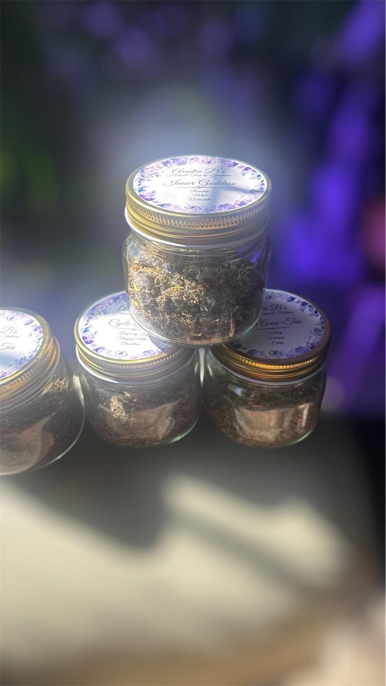 Puede incluir: Cuatro peque&ntilde;os frascos de vidrio llenos de hierbas secas, apilados en forma de pir&aacute;mide. Cada frasco tiene una tapa dorada y una etiqueta con texto y detalles florales. Los frascos est&aacute;n llenos de una mezcla de hierbas marrones y verdes.