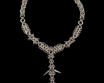 May include: A silver-toned chainmail necklace with a Y-shaped pendant. The pendant features two silver spikes and a small, off-white carved figure. The necklace is set against a black background.