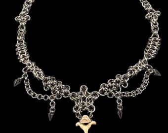 May include: Silver chainmail necklace with a bone pendant. The necklace features a series of chainmail links, decorative floral elements, and dangling silver spikes. The pendant is a light tan color.
