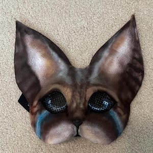 Peut inclure: Un masque en forme de chat, marron et gris, avec de grandes oreilles pointues. Le masque présente des accents bleus sous les yeux et un maillage noir couvrant les ouvertures des yeux. Accessoire de costume unique.