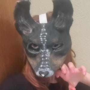 May include: A black and brown dog mask with white accents. The mask has large ears and is painted with a detailed design. The mask is worn by a person with red hair and a white bandage on their head. The person's hand is in a claw-like position.