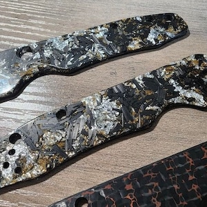 May include: Close-up of three knife handle scales with a unique pattern. The scales feature a mix of black, silver, and gold tones, creating a textured appearance. The top two scales have multiple holes for attachment. The bottom scale has a different pattern.