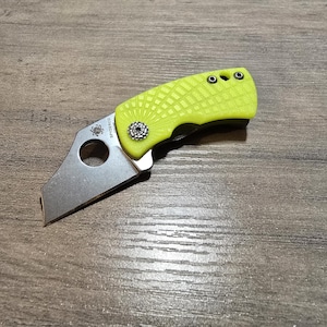May include: A small, folding utility knife with a bright yellow textured handle and a silver blade. The blade has a unique shape with a circular hole near the handle. The knife is resting on a wooden surface.