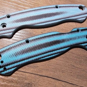 May include: Two knife handle scales with a textured pattern in shades of blue, gray, and black. The scales have multiple holes for attachment and a curved design. The background is a wooden surface.