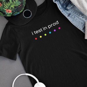 Puede incluir: Camiseta negra con el texto "i test in prod" y una serie de puntos de colores. También se ve una gorra de béisbol negra con un diseño floral y auriculares blancos.