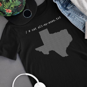 以下が含まれることがあります： 黒いTシャツに、テキサス州の白いアウトラインと「/# cat all-my-exes.txt」というテキストがプリントされています。Tシャツは、グレーの表面に、花柄の帽子と白いヘッドホンと一緒に置かれています。