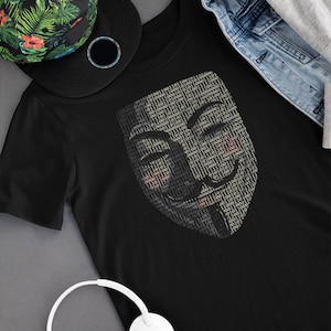 May include: A black t-shirt featuring a graphic of the Anonymous mask, created using binary code. The mask is a symbol of internet privacy and freedom of speech. The t-shirt is laid out on a grey surface with a green and black patterned hat and white headphones.