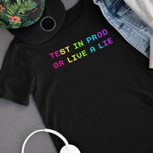 May include: Black t-shirt with the text "TEST IN PROD OR LIVE A LIE" in rainbow colors. The shirt is laid flat on a gray surface with a floral patterned hat and white headphones.