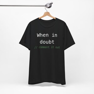Könnte beinhalten: Schwarzes T-Shirt mit dem Text "When in doubt // comment it out" in Weiß und Grün. Das T-Shirt hängt an einem Holzbügel vor weißem Hintergrund.