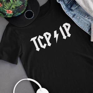 Pode incluir: Camiseta preta com texto branco que diz "ICP ⚡️ IP". A camiseta está sobre uma superfície cinza com um chapéu estampado preto e verde e um par de fones de ouvido brancos.