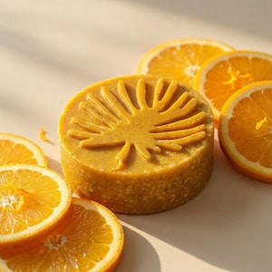 May include: A round, textured orange soap bar featuring a raised palm leaf design on its surface, surrounded by several fresh, bright orange slices arranged in a circular pattern on a neutral, light-coloured surface under soft natural lighting.