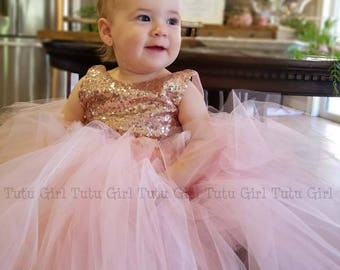 May include: A baby wearing a pink tulle dress with a gold sequined bodice. The dress has multiple layers of soft pink tulle. The text "Tutu Girl" is printed on the dress. The baby is smiling.