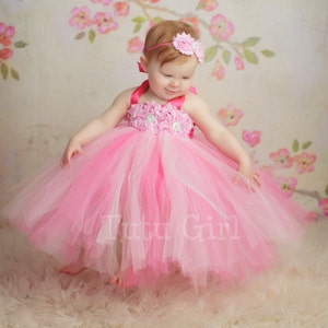 May include: A little girl wearing a pink and white tulle tutu dress with a pink flower headband. The dress has a pink satin bodice with a floral design and a pink ribbon.