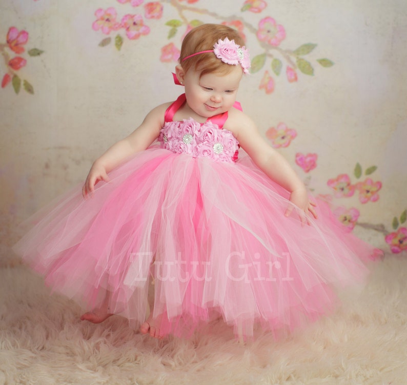 May include: A little girl wearing a pink and white tulle tutu dress with a pink flower headband. The dress has a pink satin bodice with a floral design and a pink ribbon.