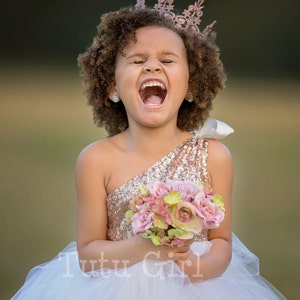 May include: A young girl wearing a white tulle dress with a gold sequined bodice and a crown. She is holding a bouquet of pink flowers and has a big smile on her face. The text "Tutu Girl" is visible on the dress.