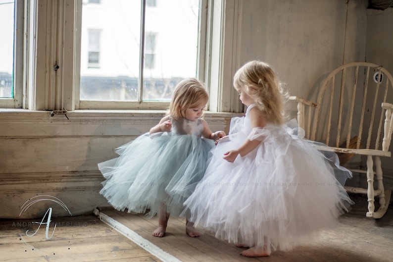 Peut inclure: Deux jeunes filles portant des robes en tulle, l'une en blanc et l'autre en bleu clair, se tiennent dans une pi&egrave;ce avec une fen&ecirc;tre et un fauteuil &agrave; bascule. Les filles se regardent et sourient.