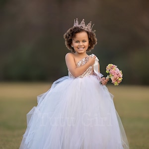 May include: A young girl wearing a white tulle dress with a gold sequined bodice and a gold crown. She is holding a bouquet of pink flowers.