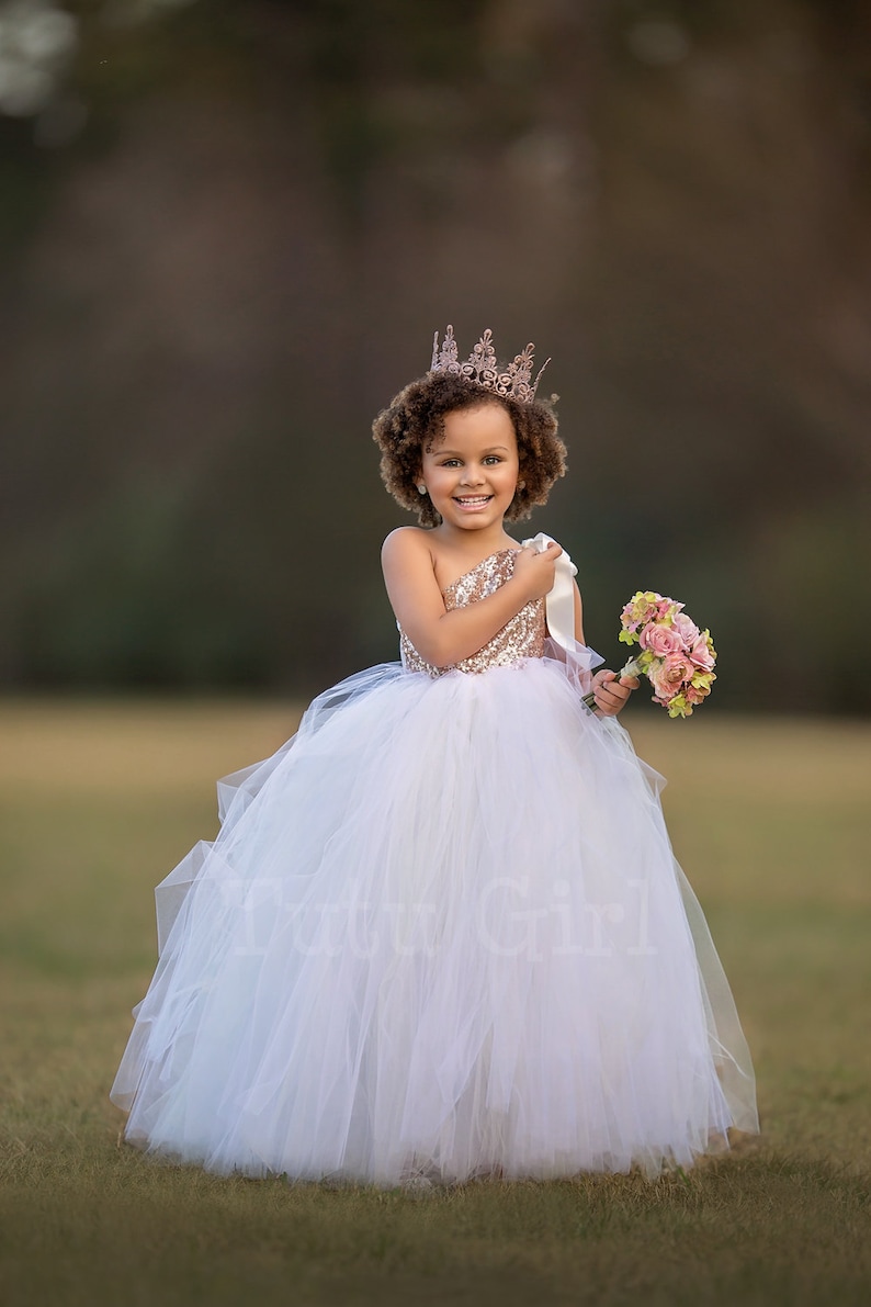 May include: A young girl wearing a white tulle dress with a gold sequined bodice and a gold crown. She is holding a bouquet of pink flowers.