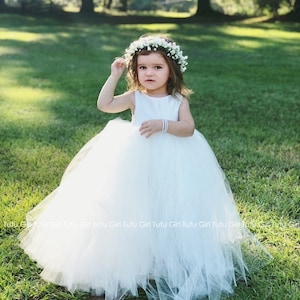 Peut inclure: Une jeune fille portant une robe en tulle blanche et une couronne de fleurs. La robe a une jupe ample et est parfaite pour une occasion sp&eacute;ciale.