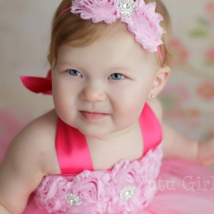 May include: A baby girl wearing a pink tulle dress with a pink flower headband and a pink bow. The dress has a pink satin ribbon and a rhinestone embellishment. The headband has a rhinestone center.