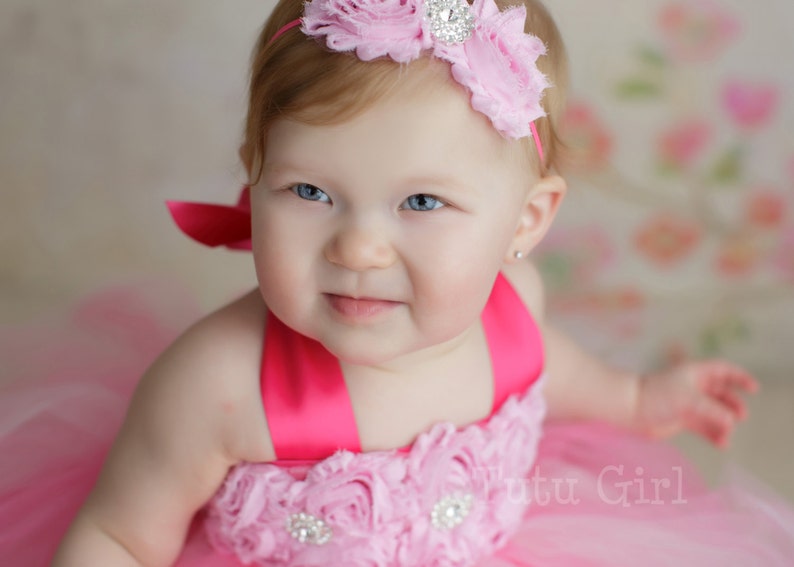 May include: A baby girl wearing a pink tulle dress with a pink flower headband and a pink bow. The dress has a pink satin ribbon and a rhinestone embellishment. The headband has a rhinestone center.