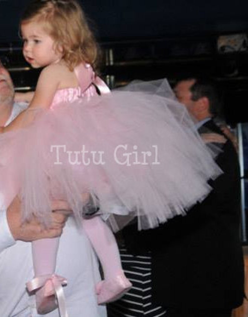 Pu&ograve; includere: Una bambina che indossa un tut&ugrave; di tulle rosa con un corpetto di raso bianco e un fiocco bianco. Il tut&ugrave; &egrave; molto pieno e soffice. La bambina sorride e guarda la fotocamera. Il testo "Tutu Girl" &egrave; stampato sul tut&ugrave;.