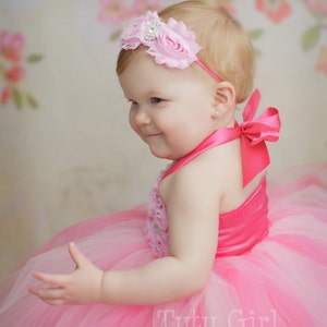 May include: A baby girl wearing a pink tutu dress with a pink flower headband and a pink bow on the back of the dress. The dress has a pink satin bodice with a floral design. The background is a floral pattern.