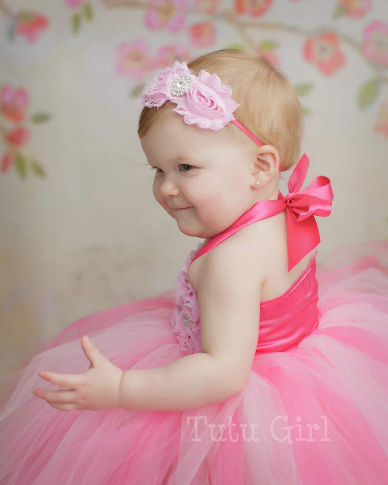 May include: A baby girl wearing a pink tutu dress with a pink flower headband and a pink bow on the back of the dress. The dress has a pink satin bodice with a floral design. The background is a floral pattern.
