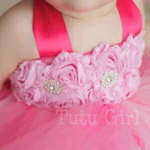 May include: A pink tulle tutu dress with a pink and white flower embellishment and a pink satin ribbon. The dress has a rhinestone embellishment in the center of the flower.
