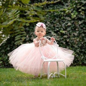 May include: A young girl wearing a pink tulle dress with gold sequins and a pink bow headband. She is standing next to a white metal chair in a grassy area.