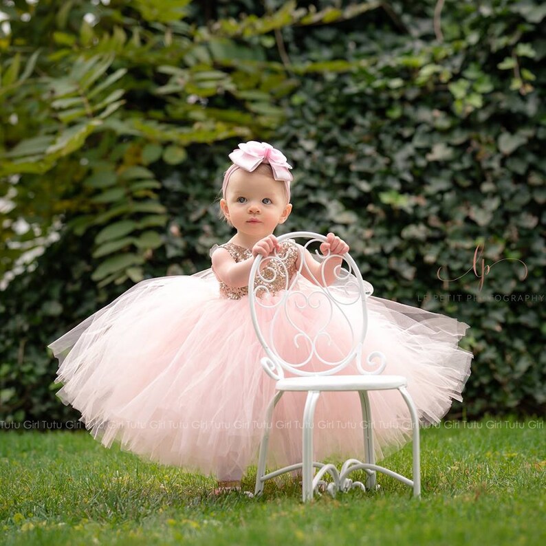 May include: A young girl wearing a pink tulle dress with gold sequins and a pink bow headband. She is standing next to a white metal chair in a grassy area.
