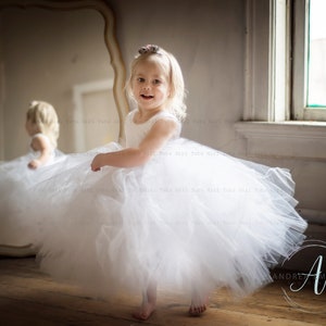 Peut inclure: Une jeune fille v&ecirc;tue d'une robe en tulle blanche avec un bandeau de fleurs sourit &agrave; la cam&eacute;ra. Elle se tient devant un miroir et une fen&ecirc;tre.