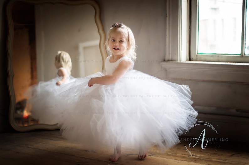 Peut inclure: Une jeune fille v&ecirc;tue d'une robe en tulle blanche avec un bandeau de fleurs sourit &agrave; la cam&eacute;ra. Elle se tient devant un miroir et une fen&ecirc;tre.