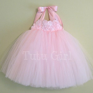 Pu&ograve; includere: Un abito tutu di tulle rosa con un top halter in nastro di raso rosa e un ricamo di fiori intorno al corpetto. L'abito ha il testo "Tutu Girl" stampato sulla gonna.