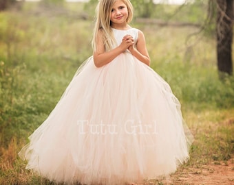 May include: A young girl wearing a white sleeveless bodice and a blush pink tulle tutu skirt. The skirt is very full and has multiple layers. The girl is standing on a dirt path with green grass and trees in the background. The text "Tutu Girl" is visible on the skirt.