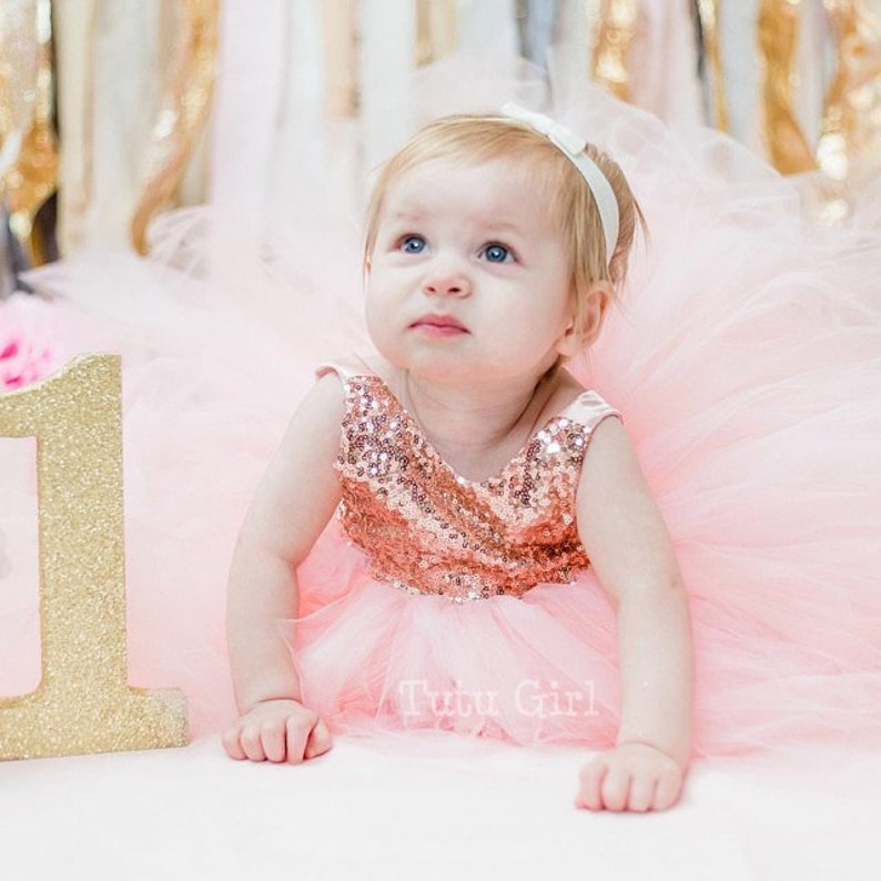 May include: A baby girl wearing a pink tulle dress with rose gold sequins and a white headband. The dress has a tulle skirt and a sequined bodice. The baby is sitting on a pink surface with a gold glitter number one.