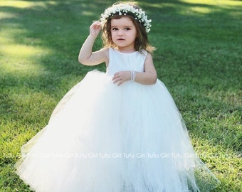 May include: A young child in a light ivory, off-white, sleeveless tutu dress and a floral crown. The dress features a full, layered skirt. The child is standing on green grass in a sunny outdoor setting.