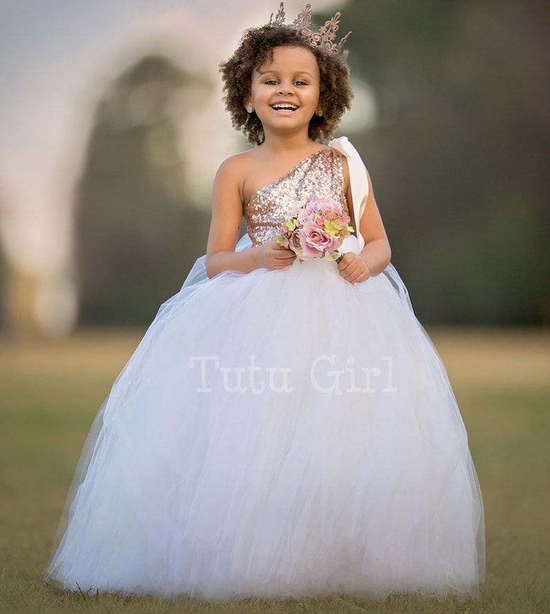 May include: A young girl wearing a white tulle dress with a rose gold sequin top and a white sash. She is holding a bouquet of pink flowers and wearing a gold crown. The dress is labeled "Tutu Girl".
