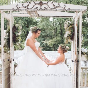 May include: A bride and a flower girl in white tulle dresses stand under a white wooden archway decorated with greenery. The bride is wearing a flower crown and the flower girl is holding a bouquet of flowers. The text "Rose Gold Sequins Off-White Skirt" is visible at the bottom of the image.