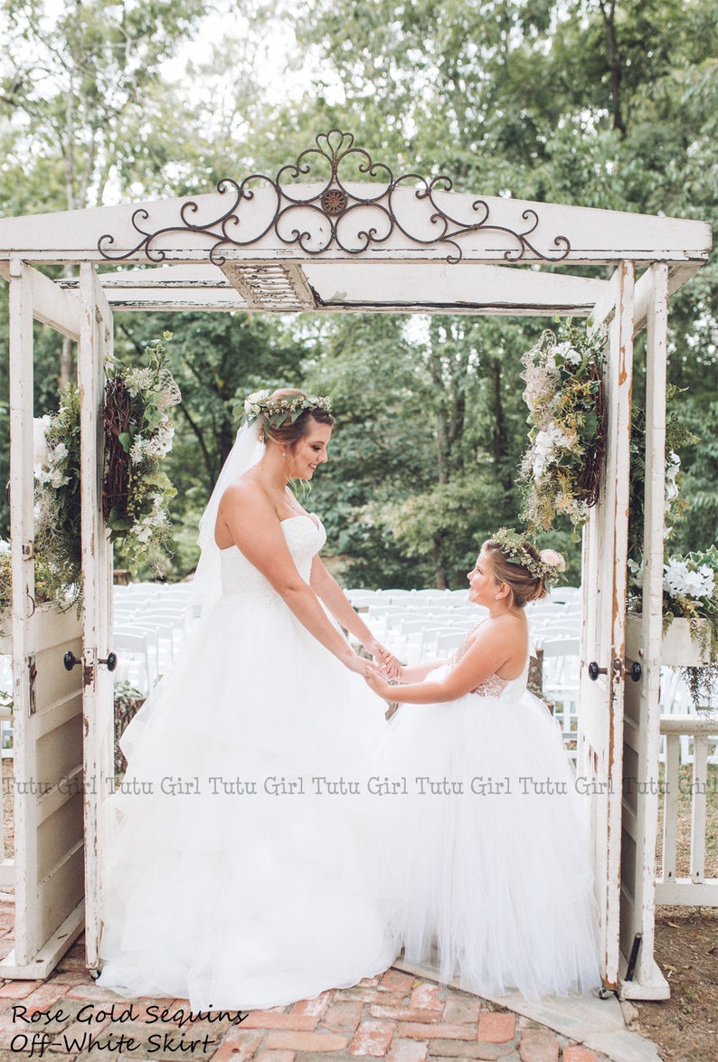 May include: A bride and a flower girl in white tulle dresses stand under a white wooden archway decorated with greenery. The bride is wearing a flower crown and the flower girl is holding a bouquet of flowers. The text "Rose Gold Sequins Off-White Skirt" is visible at the bottom of the image.
