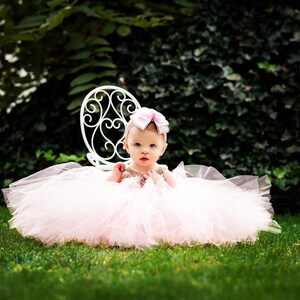 May include: A baby girl wearing a pink tulle dress and a pink bow headband. She is sitting on a white metal chair in a grassy area.