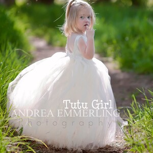 Peut inclure: Une jeune fille portant une robe en tulle blanche et un bandeau blanc, debout sur un chemin dans une zone herbeuse. Le texte "Tutu Girl Andrea Emmerling Photography" est visible sur la robe.