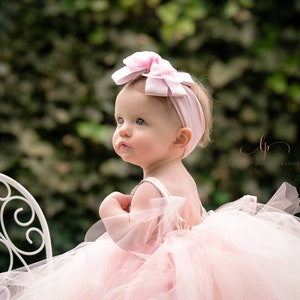 May include: A baby girl wearing a pink tulle tutu dress and a pink bow headband. She is sitting on a white chair and looking up.