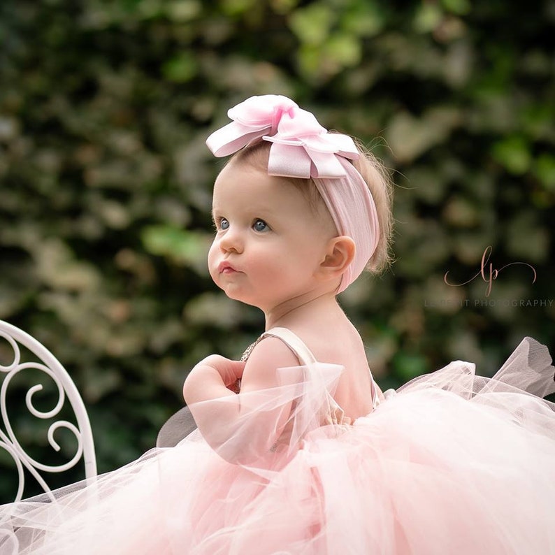 May include: A baby girl wearing a pink tulle tutu dress and a pink bow headband. She is sitting on a white chair and looking up.