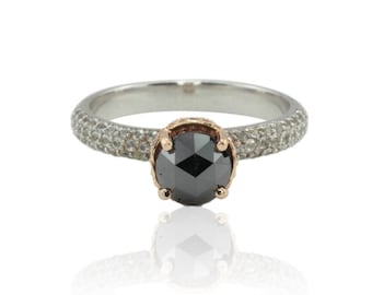 May include: A silver ring with a black diamond center stone. The band is encrusted with small, clear stones. The diamond is set in a rose gold bezel. The ring is displayed on a white background.