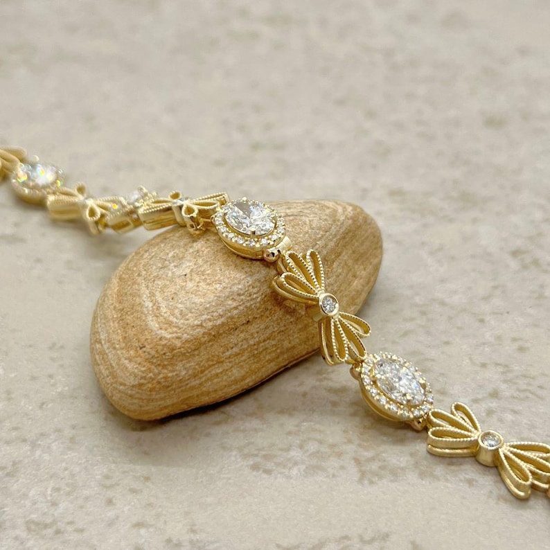 May include: A gold bracelet with a delicate bow design and oval-shaped clear stones. The bracelet is on a light brown rock.