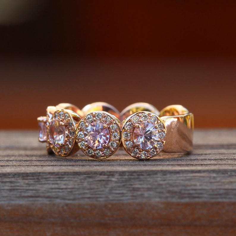 Puede incluir: Un anillo de oro rosa con varias piedras preciosas rosas. El anillo presenta una serie de piedras rosas redondas engastadas en halo, cada una rodeada de piedras m&aacute;s peque&ntilde;as y transparentes. La banda es de oro rosa pulido, sobre una superficie de madera.