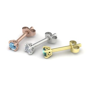 May include: Three stud earrings in rose gold, silver, and gold. Each earring features a single gemstone in a four-prong setting. The gemstones are blue, clear, and teal. The earrings are displayed on a white background.